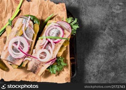 Dutch sandwich with herring, pickled cucumbers and red onions on a paper lining. Ingredients on a cutting board nearby. Layout on the table, selective focus and copy space