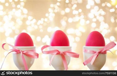 easter and holidays concept - pink colored eggs in ceramic cup holders with ribbon over festive lights on beige background. pink easter eggs in holders over festive lights