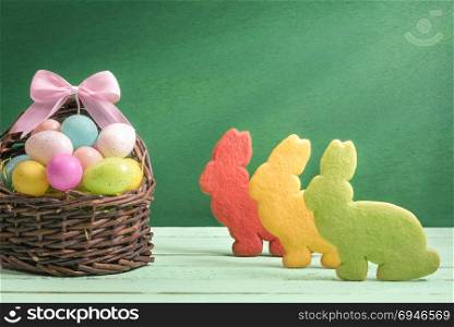 Easter banner design with a wicker basket full of colorful painted eggs, with a ribbon bow and three bunny shaped cookies, on a green background.