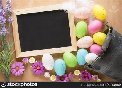 Easter card with a rustic metal bucket full of colorful painted eggs, overturned on the table and an unwritten blackboard surrounded by flowers.