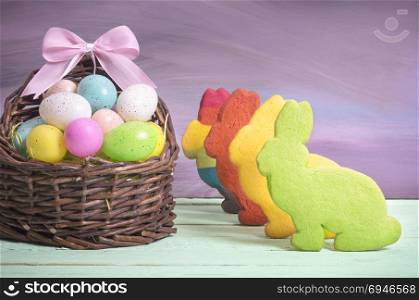 Easter card with a wicker basket full of painted eggs, with a pink ribbon bow and multicolor bunny shaped cookies, on a green table and purple wall.
