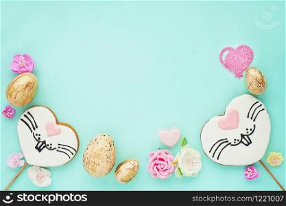 Easter composition on a blue background. Gingerbread cookies with a painted rabbit face, golden eggs, paper flowers and hearts. Flat layout, top view, space for copy.