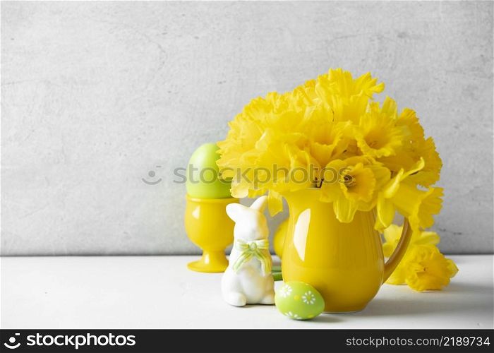 Easter composition with Easter eggs, bunny and Bouquet of yellow daffodils in a yellow jug on table top, home decor, interior, copy space