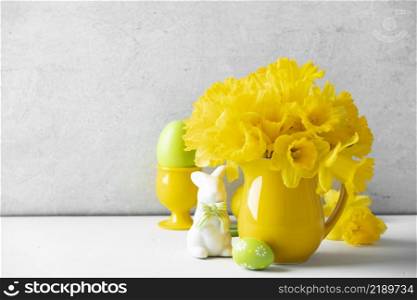 Easter composition with Easter eggs, bunny and Bouquet of yellow daffodils in a yellow jug on table top, home decor, interior, copy space