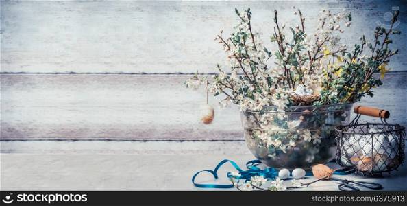 Easter concept with springtime decoration setting : bunch of spring branches with cherry blossom, basket with eggs, ribbon and scissors on table at wall background, front view, banner