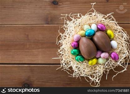 easter, confectionery and holidays concept - chocolate eggs and candies in straw nest on wooden background. chocolate eggs and candies in straw nest