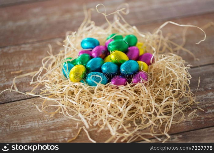 easter, confectionery and holidays concept - chocolate eggs in foil wrappers in straw nest on wooden table. chocolate eggs in foil wrappers in straw nest