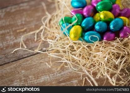 easter, confectionery and holidays concept - chocolate eggs in foil wrappers in straw nest on wooden table. chocolate eggs in foil wrappers in straw nest