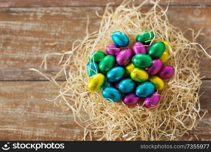 easter, confectionery and holidays concept - chocolate eggs in foil wrappers in straw nest on wooden background. chocolate eggs in foil wrappers in straw nest