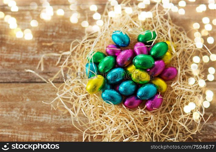 easter, confectionery and holidays concept - chocolate eggs in foil wrappers in straw nest on wooden background. chocolate eggs in foil wrappers in straw nest