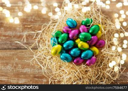 easter, confectionery and holidays concept - chocolate eggs in foil wrappers in straw nest on wooden background. chocolate eggs in foil wrappers in straw nest