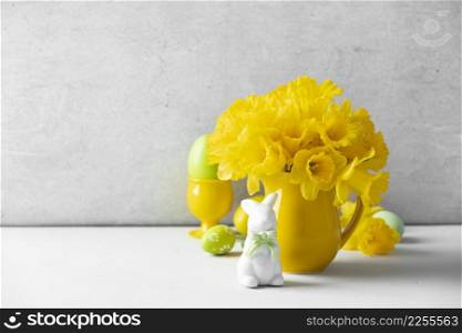 Easter eggs, bunny and Bouquet of yellow daffodils in a yellow jug on table top, Easter composition, home decor, interior, copy space
