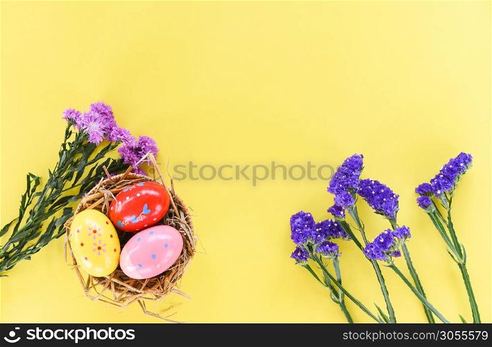 Easter eggs in basket nest decoration with purple flower Marguerite and statice flowers on yellow background - top view