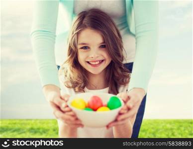 easter, family, holiday and child concept - smiling little girl and mother holding bowl with colored eggs
