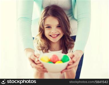 easter, family, holiday and child concept - smiling little girl and mother holding bowl with colored eggs