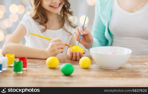 easter, family, holiday, people and childhood concept - close up of happy girl and mother with brushes coloring easter eggs over lights background