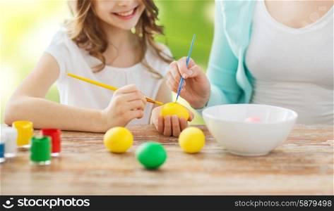 easter, family, holiday, people and childhood concept - close up of happy girl and mother with brushes coloring easter eggs over lights background