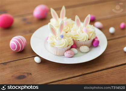 easter, food and holidays concept - frosted cupcakes with colored eggs and candies on wooden table. cupcakes with easter eggs and candies on table