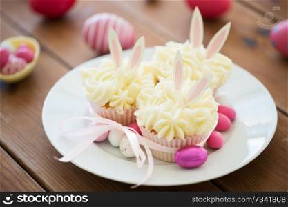 easter, food and holidays concept - frosted cupcakes with colored eggs and candies on wooden table. cupcakes with easter eggs and candies on table