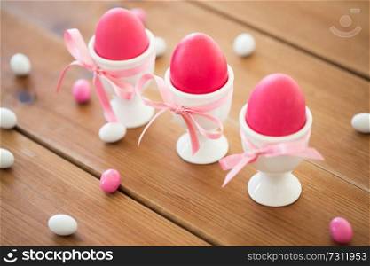 easter, food and holidays concept - pink colored eggs in ceramic cup holders with ribbon and candy drops on wooden table. easter eggs in holders and candy drops on table