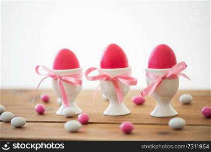 easter, food and holidays concept - pink colored eggs in ceramic cup holders with ribbon and candy drops on wooden table. easter eggs in holders and candy drops on table