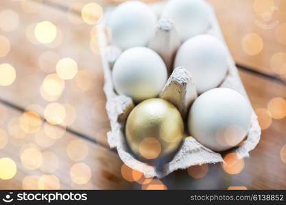 easter, food, cooking and object concept - close up of white and golden eggs in egg box or carton wooden surface over holidays lights