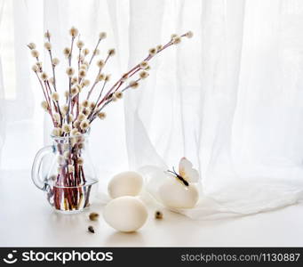 Easter greeting card. Fluffy pussy-willow branches in a transparent glass pitcher and three white Easter eggs against a white transparent cloth, with copy-space