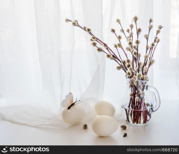 Easter greeting card. Fluffy pussy-willow branches in a transparent glass pitcher and three white Easter eggs against a white transparent cloth