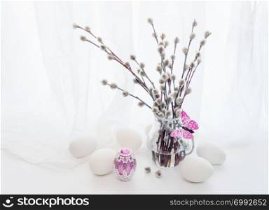 Easter greeting card. Fluffy pussy-willow branches in a transparent glass pitcher, pink batterfly and white end pink Easter eggs against a white transparent cloth