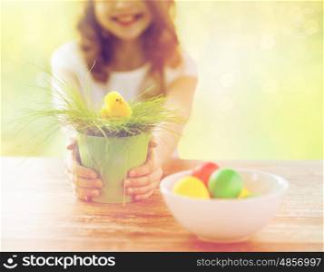 easter, holiday and child concept - close up of girl holding pot with green grass and yellow chicken toy with bowl of colored eggs over green background