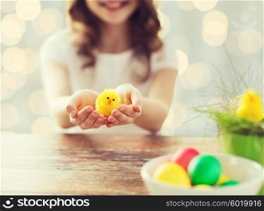 easter, holiday and child concept - close up of girl holding pot with green grass, yellow chicken toy and bowl of colored eggs on table over lights background