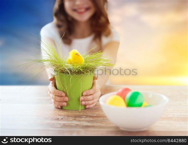 easter, holiday and child concept - close up of girl holding pot with green grass and yellow toy chicken and bowl of colored eggs on table over sky background. close up of girl with easter toy chicken and eggs