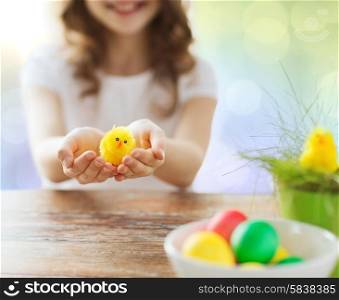 easter, holiday and child concept - close up of girl holding yellow chicken toy and bowl of colored eggs on table over lights background
