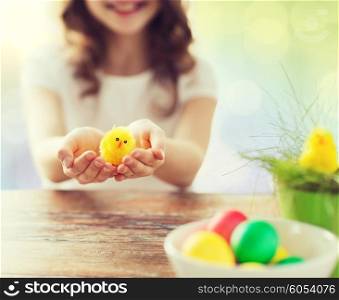 easter, holiday and child concept - close up of girl holding yellow chicken toy and bowl of colored eggs on table over lights background