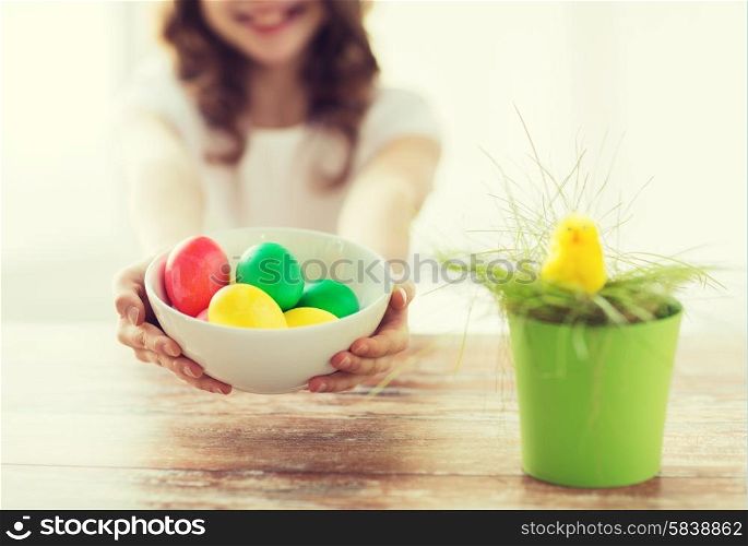 easter, holiday and child concept - close up of little girl holding bowl with colored eggs and green grass in pot with chicken