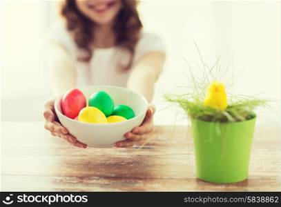 easter, holiday and child concept - close up of little girl holding bowl with colored eggs and green grass in pot with chicken