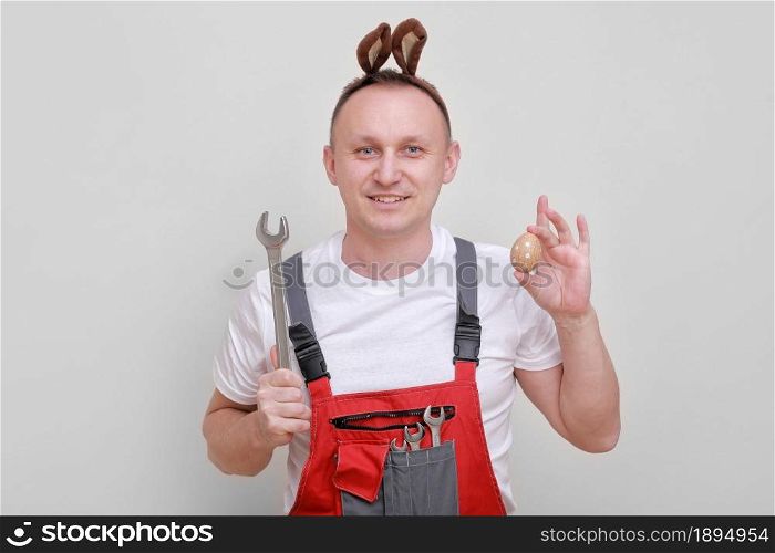 Easter holiday, celebration concept. funny smiling engineer worker or mechanic is wearing rabbit ears on head, holding wrenches and painted egg white background. celebrating orthodox day.. Easter holiday, celebration concept. funny smiling engineer worker or mechanic is wearing rabbit ears on head, holding wrenches and painted egg white background. celebrating orthodox day