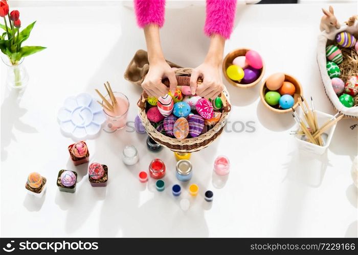 Easter holiday concept, Close up of hand of holding wearing bunny ears hand holding a basket with colorful Easter eggs on white wooden table background in top view
