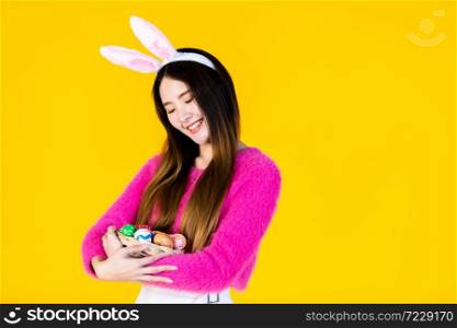 Easter holiday concept, Happy Asian Young woman wearing bunny ears hand holding a basket with colorful Easter eggs In isolated on Yellow blank copy space studio background.