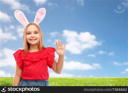 easter, holidays and childhood concept - happy girl wearing bunny ears headband waving hand over blue sky and grass background. happy girl wearing easter bunny ears waving hand