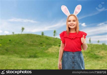 easter, holidays and childhood concept - happy girl wearing bunny ears headband showing thumbs up over blue sky and green meadow background. girl wearing easter bunny ears showing thumbs up