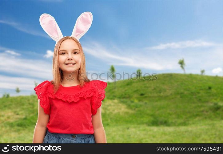 easter, holidays and childhood concept - happy girl wearing bunny ears headband over blue sky and green meadow background. happy girl wearing easter bunny ears headband