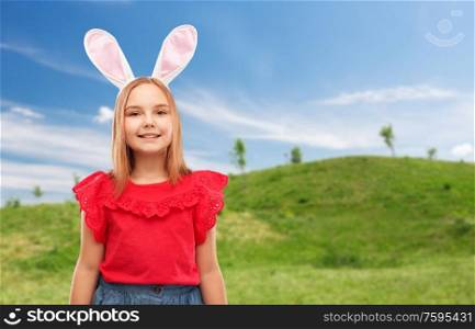 easter, holidays and childhood concept - happy girl wearing bunny ears headband over blue sky and green meadow background. happy girl wearing easter bunny ears headband