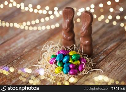 easter, holidays and object concept - close up of candy eggs in straw nest and chocolate bunnies on wooden table. easter eggs in straw nest and chocolate bunnies