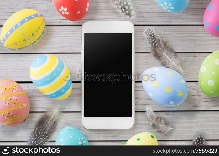 easter, holidays and object concept - close up of colored eggs and feathers over grey wooden boards background. smartphone with easter eggs and feathers