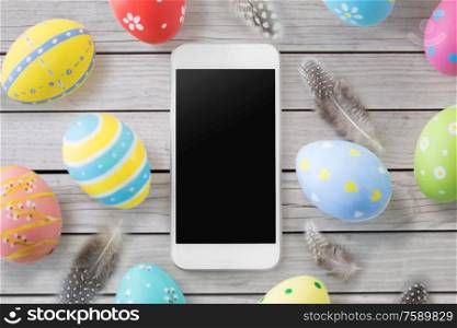 easter, holidays and object concept - close up of colored eggs and feathers over grey wooden boards background. smartphone with easter eggs and feathers