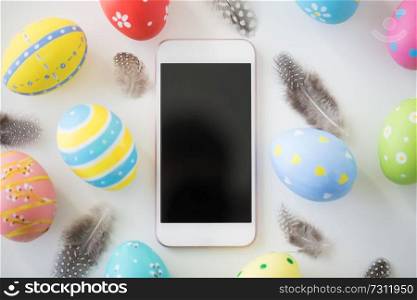 easter, holidays and object concept - close up of colored eggs and feathers on white background. smartphone with easter eggs and feathers