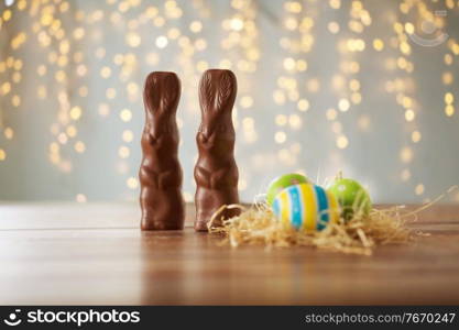 easter, holidays and object concept - close up of colored eggs in straw nest and chocolate bunnies on wooden table over bokeh lighs. easter eggs in straw nest and chocolate bunnies