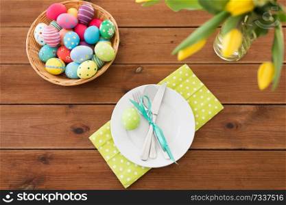 easter, holidays and object concept - colored eggs in basket, plates, cutlery and flowers on wooden table. easter eggs in basket, plates, cutlery and flowers