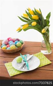 easter, holidays and object concept - colored eggs in basket, plates, cutlery and flowers on wooden table. easter eggs in basket, plates, cutlery and flowers